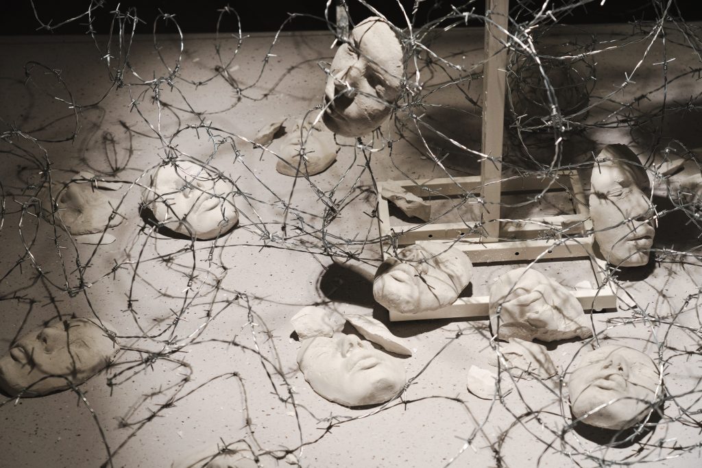 DSC000419 Horizon, 2025: plaster casts of the artist's face lying among loops of barbed wire on a pale surface, a wooden frame standing behind them.