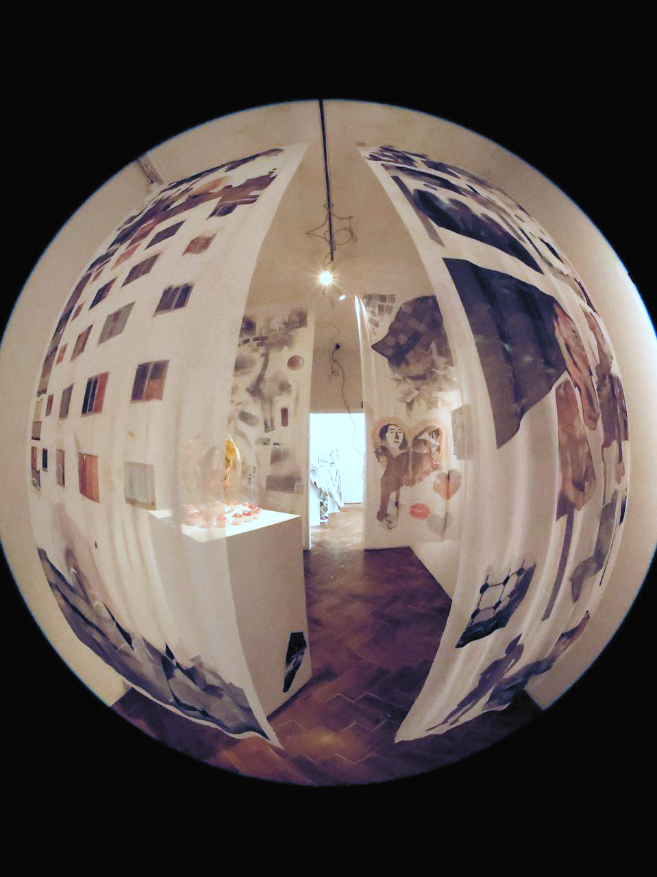 The exhibition through a fisheye lens: the whole room bent into a circle floating on black — printed panels hanging from the ceiling, a lit plinth, a bare bulb and a doorway through to the next room.